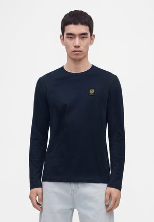 Junger Mann trägt ein schwarzes Langarmshirt mit kleinem Brustlogo und hellblauen Jeans und steht vor einem einfarbigen Hintergrund.
