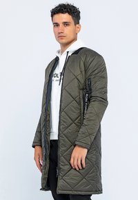 Olivengrüner gesteppter langer Mantel mit einem Reißverschluss, der mit einem schwarzen Hardware-Akzent versehen ist und einer weißen Kapuzenjacke, die darunter getragen wird.