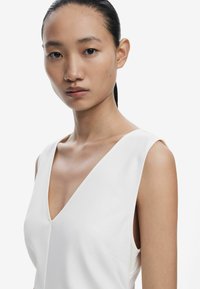 Rochie albă fără mâneci, cu decolteu adânc în V, textură de catifea fină și cusături minime. Modelul are o postură dreaptă și părul lins îndback.