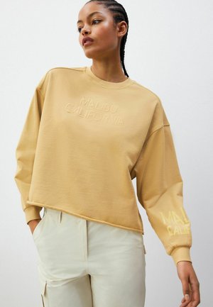 Beige cropped sweatshirt with embroidered "MALIBU CALIFORNIA" in matching color, featuring drop shoulders and long sleeves.