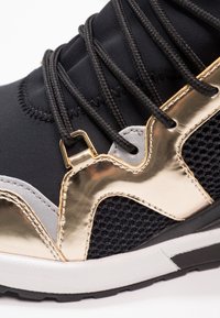 Close-up of black sneaker with gold metallic accents, black mesh, thick black laces, and white sole with black tread pattern.