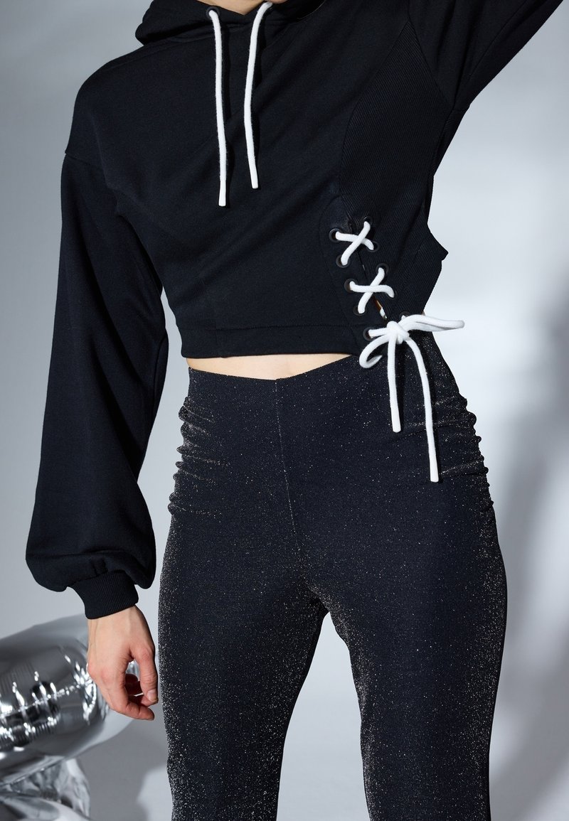 Černá zkrácená mikina s bílými šňůrkami a šněrováním po stranách, doplněná vysokými černými legínami s lesklou texturou.
