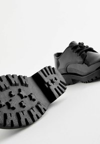 Chaussures noires en cuir verni avec un bout arrondi, à lacets, et semelle en caoutchouc texturé dotée de motifs d'étoiles et de rainures de traction.