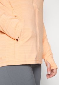 Jaqueta com fecho zip em tom pêssego, com textura horizontal, apresentando um bolso lateral e buracos para os polegares nas mangas. Usada com calças cinzas.