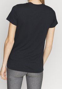 Person som bär en enkel svart t-shirt med kort ärm och gråa tights, sedd från ryggen mot en enfärgad ljus bakgrund.