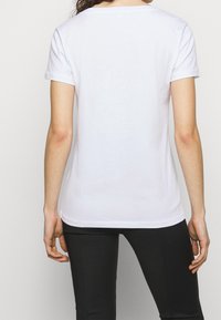 Bílá bavlněná tričko s krátkými rukávy, kulatým výstřihem a rovým lemem. Látka vypadá hladce a lehce, bez viditelných vzorů.