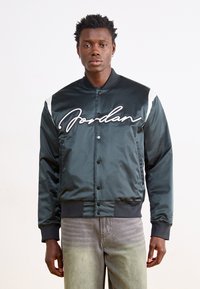 Grønn sateng bomberjakke med hvite accentstriper og brodert "Jordan"-logo. Har trykknap-lukking og ribbestrikkede mansjetter.