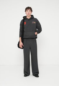 Γκρι φούτερ με κουκούλα και τσέπη μπροστά, με πορτοκαλί και λευκό λογότυπο· συνδυασμένο με γκρι παντελόνι φαρδιάς γραμμής. Το μοντέλο κρατά μια πλεκτή μαύρη τσάντα.