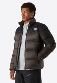 Hnědá a černá bundy s kapucí s vysokým límcem, zipem a logy. Má prošívanou texturu a boční kapsy. Nosená přes šedou mikinu.