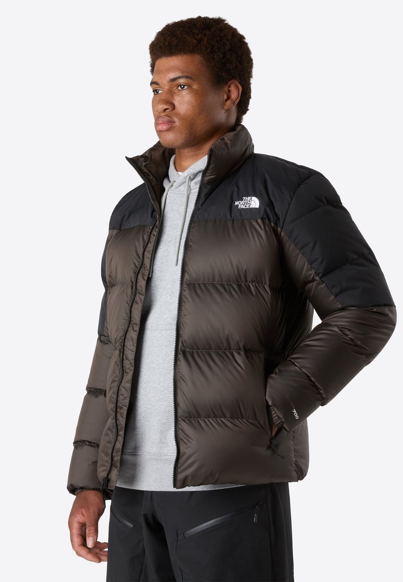 Braune und schwarze Steppjacke mit hohem Kragen, Reißverschluss und Logos. Verfügt über eine gesteppten Textur und seitliche Taschen. Getragen über einem grauen Hoodie.