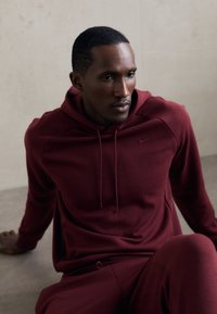 Męski zestaw w kolorze bordowym składający się z bluzy z kapturem i spodni dresowych, wykonany z miękkiego materiału, z kapturem na sznurki i subtelnym logo Nike. Gładka tekstura, sportowy krój.