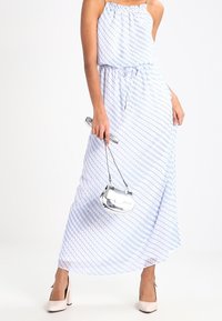 Donna che indossa un vestito maxi senza maniche bianco e azzurro a fantasia, mentre tiene una piccola borsa argentata, abbinata a tacchi alti di un pallido rosa.