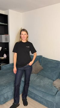 Γυναίκα που χαμογελά με το χέρι στην τσέπη, φοράει μαύρο t-shirt AllSaints, σκουρόχρωμο τζιν και μαύρα παπούτσια, στέκεται δίπλα σε έναν μπλε καναπέ σε ένα σαλόνι.