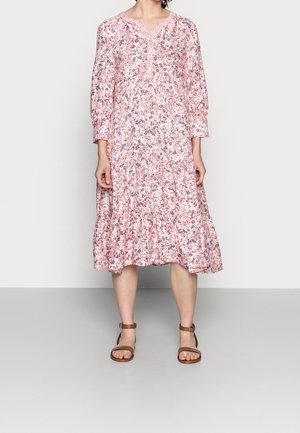 Femme portant une robe midi ample rose à fleurs avec des manches longues et des sandales marron à bride cheville, debout devant un fond uni.