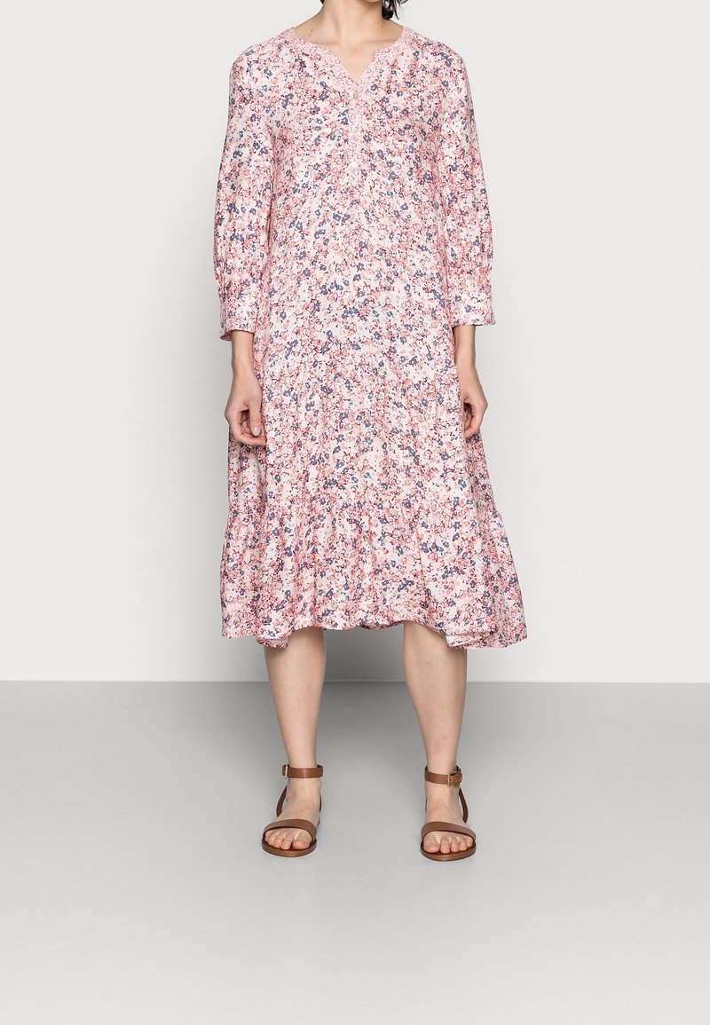 Femme portant une robe midi ample rose à fleurs avec des manches longues et des sandales marron à bride cheville, debout devant un fond uni.