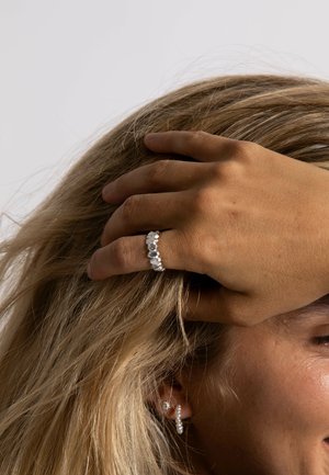 Silver ring with multiple clear gemstones, worn on a hand touching blonde hair. Accompanied by pearl and diamond stud earrings.