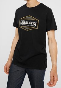 Svart bomull T-shirt med en stor gyllene hexagonal Billabong-logotyp och texten "SINCINETEEN SEVENTY THREE", korta ärmar och avslappnad passform.