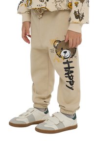 Beżowe spodnie dresowe z grafiką niedźwiedzia i czarnym napisem "HAPPY". Elastyczny pas, ściągacze u kostek, miękka tekstura. Noszone z białymi trampkami.