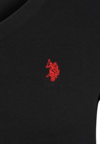 Pull en maille noire avec un design en col en V et un petit logo rouge brodé sur la poitrine. La texture est lisse et légèrement côtelée.