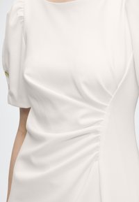 Blouse blanche à manches courtes avec un détail froncé sur un côté et un petit bouton doré sur la manche.