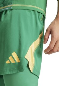Pantaloni sportivi verdi realizzati in materiale leggero, con un accento giallo sul lato e un logo Adidas giallo nella parte inferiore sinistra.