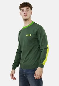 Maglione verde a maniche lunghe con collo verde brillante, dettagli gialli sulle maniche e un logo minimale giallo sul petto. Texture morbida.