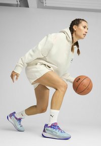 Sudadera con capucha de color claro y pantalones cortos, zapatillas deportivas azules y moradas con un diseño texturizado, sosteniendo un balón de baloncesto.