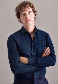Marineblaues Button-Down-Hemd mit langen Ärmeln, glatter Textur und kontrastierendem gemustertem Kragen. Das Model steht mit verschränkten Armen vor einem neutralen Hintergrund.