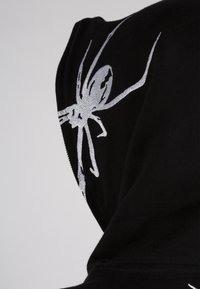 Sudadera negra con capucha que presenta un diseño de araña plateada en la capucha. Material que parece suave con una textura lisa; corte holgado.