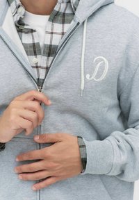 Personne en train de fermer une veste à capuche gris clair avec un logo "D" blanc, portant une chemise à carreaux blanche et grise en dessous, les mains en pleine action.