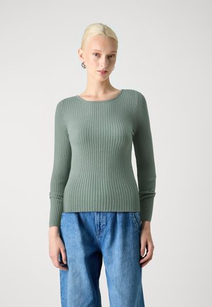 Green ribbed knit sweater with long sleeves, rounded neckline, and rolled cuffs, paired with blue loose-fitting denim pants.