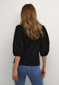 Blusa negra acolchada con mangas abullonadas, tejido texturizado, dobladillo redondeado y puños elásticos. Combinada con jeans de mezclilla azules.