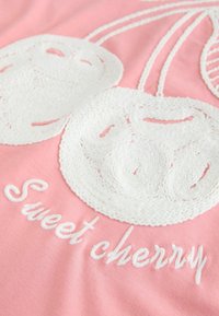 Rožnata tkanina z belim vezenjem češenj s spiralnim vzorcem, spremljana z napisom "Sladka češnja" v belem sukancu.