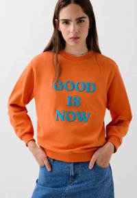 Felpa arancione con testo ricamato blu "GOOD IS NOW." Presenta un collo tondo e polsini a coste, abbinata a una gonna di jeans.