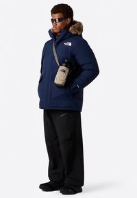 Tmavě modrá zateplená parka s kapucí lemovanou kožešinou, doplněná černými kalhotami a černými botami, s béžovou taškou na vodní láhev.