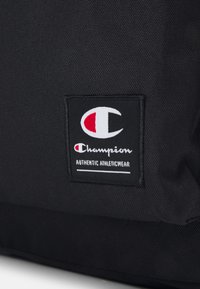 Sac en tissu noir avec un écusson logé rectangulaire de Champion avec des détails rouges et blancs, étiqueté "AUTHENTIC ATHLETICWEAR." Surface texturée.