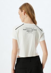 Witte cropped t-shirt met zwarte accentstiksels, met op de achterkant de tekst: "WINS IT / DREAM IT WANT IT / GO FOR IT / RACEVIBES."