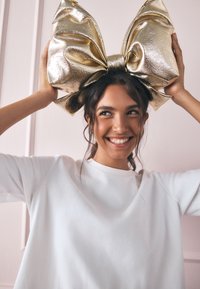 Kultainen metallinhohtoinen suurempi rusettipanta, kiiltävä pinta, sijoitettu päähän. Malli käyttää yksinkertaista valkoista collegepaitaa. Vaaleanpunainen taustaseinä.