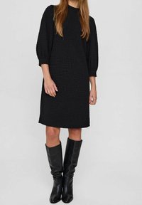 Svart knälång klänning med en texturerad tyg, rund halsringning och puffärmar, i kombination med knähöga svarta läderstövlar.