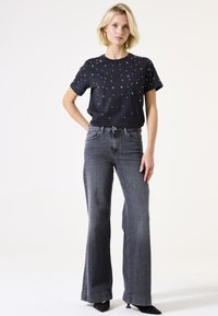 Marineblaues T-Shirt mit silbernen Sternen, kombiniert mit hochtaillierten, dunkelgrauen Schlagjeans. Das Modell steht selbstbewusst mit den Händen in den Hüften.