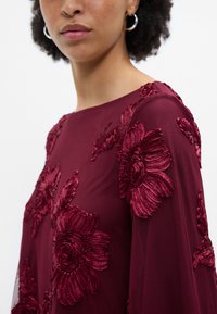 Βουργουνδίζα μπλούζα με ανάγλυφη floral κεντητική σ�ε υφή υφάσματος, με στρογγυλή λαιμόκοψη και μακριά μανίκια, απεικονιζόμενη από το πλάι.