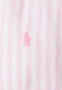 Randig tyg i vitt och ljusrosa, med en liten broderad logotyp. Materialet verkar ha en struktur som liknar linne.