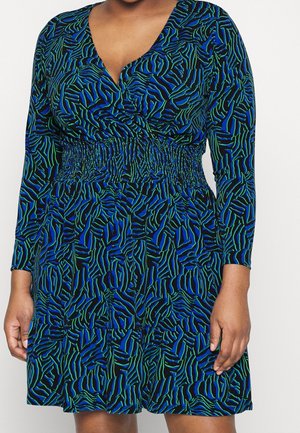 Vrouw draagt een blauwe, zwarte en groene dierenprintjurk met lange mouwen, een V-hals en een gerimpelde taille, staand tegen een witte achtergrond.