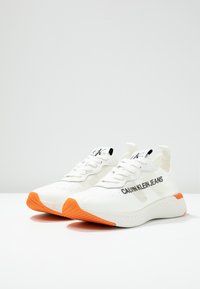 White Calvin Klein Jeans sneakers with orange soles, black logo on sides and tongue, and pull tabs at heels, shown side by side on white surface.
