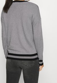 Nainen, joka on pukeutunut harmaaseen neulepuseroon, jossa on mustat ja harmaat raidalliset hihansuut ja helma, sekä tummanharmaisiin farkkuihin, seisoo valkoista taustaa vasten.