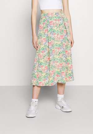 Woman wearing a colorful floral midi skirt paired with white sneakers and a white cropped top against a plain background.