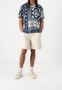 Camicia a maniche corte con un motivo floreale blu, bianco e verde; abbinata a pantaloni corti in corduroy color crema e sneakers beige.