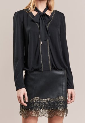 Kobieta ubrana w czarną, długorękawową bluzkę z kokardą przy szyi oraz czarną skórzaną spódnicę ozdobioną złotą koronką na dole.