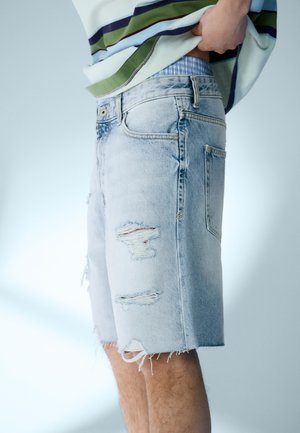 Szemüveges személy világoskék, szakadt farmer rövidnadrágot visel, amely alatt kék csíkos fehérnemű látható, valamint színes csíkos pólót, és kissé felemelik.