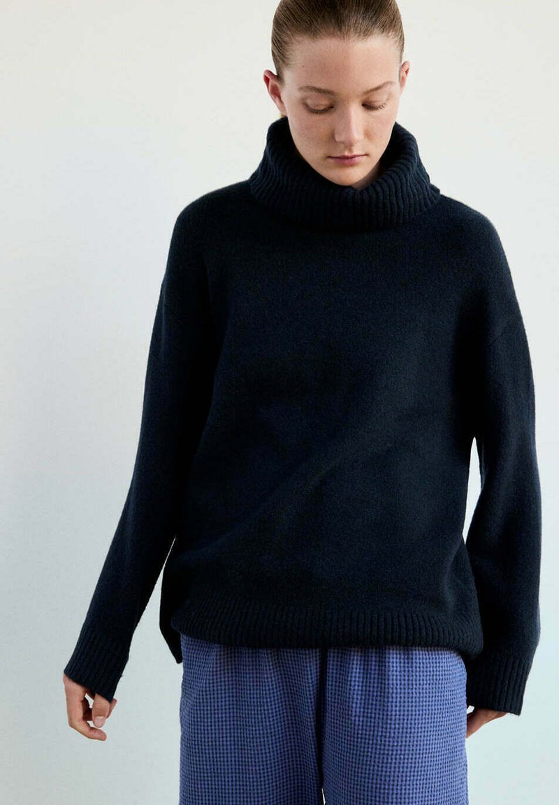 Femme à la peau claire et aux cheveux attachés, portant un pull à col roulé noir et un pantalon bleu texturé, regardant vers le bas avec une expression neutre.
